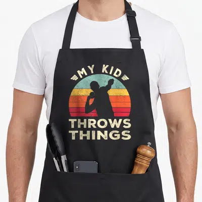Vintage field track apron - Retro sunburst with grey thrower silhouette and text My Kid Throws Things — shown on a black cotton canvas apron against a white background