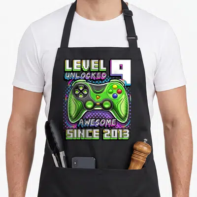 Unlocked 9th birthday video game apron - Green game controller with LEVEL UNLOCKED 9 text and colorful dots — shown on a black cotton canvas apron against a white background