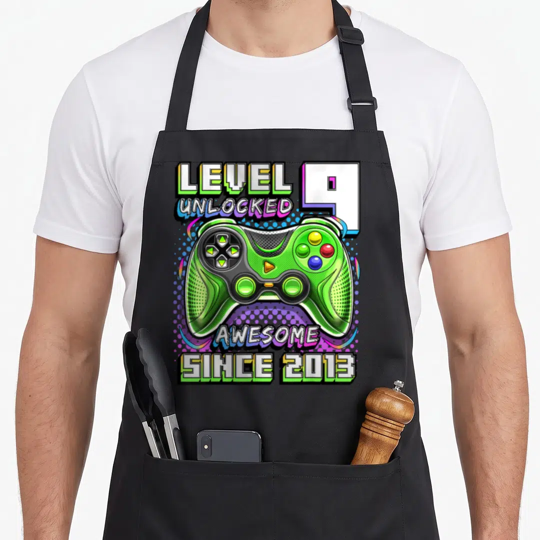 Unlocked 9th birthday video game apron - Green game controller with LEVEL UNLOCKED 9 text and colorful dots — shown on a black cotton canvas apron against a white background