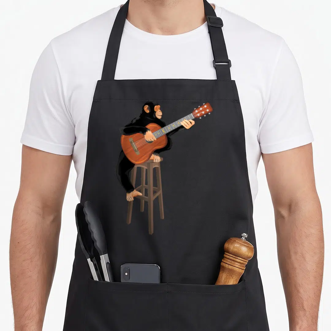Chimpanzee acoustics apron - Cartoon chimpanzee sitting on a stool playing an acoustic guitar in black, brown, and tan — shown on a black cotton canvas apron against a white background