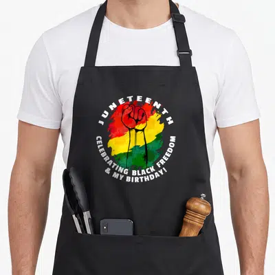 Juneteenth birthday apron - Raised fist over red yellow green splash with circular white text celebrating black freedom — shown on a black cotton canvas apron against a white background