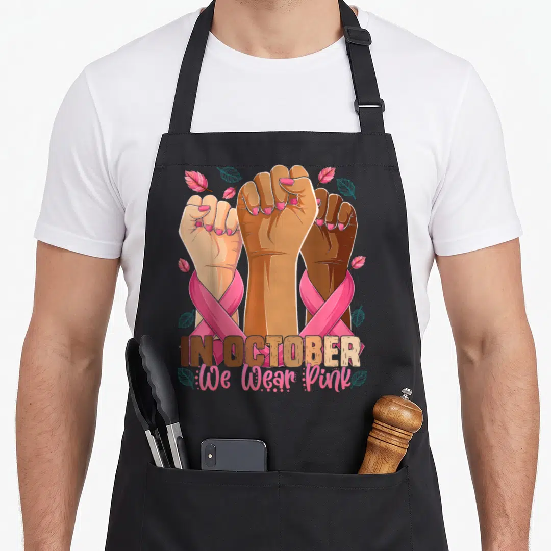 Month breast cancer awareness aprons - Three raised fists in tan brown and blue wrapped in pink ribbons with scattered — shown on a black cotton canvas apron against a white background