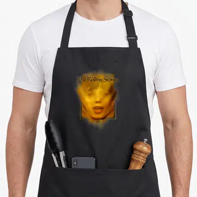 Soup rolling stones apron - Man's face in gold photo style with Rolling Stones text and Goats Head Soup caption inside — shown on a black cotton canvas apron against a white background