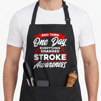 Changed stroke awareness apron - Red ribbon flanking vertical text reading And Then One Day Everything Changed Stroke — shown on a black cotton canvas apron against a white background