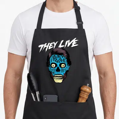 They face apron - Blue alien skull with THEY LIVE text in white and dark purple cartoon style — shown on a black cotton canvas apron against a white background