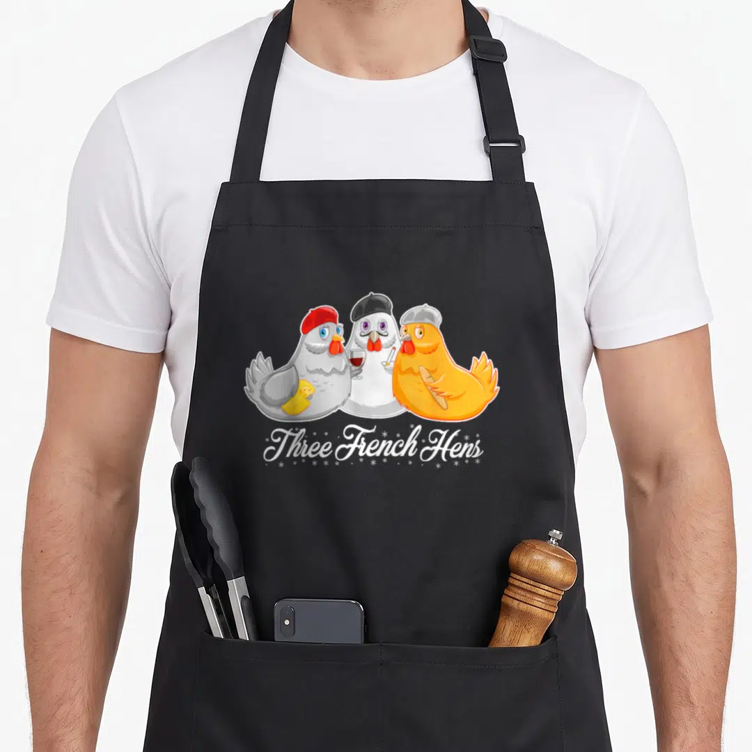 Three christmas apron - Three cartoon hens in berets holding cheese wine and bread with text Three French Hens — shown on a black cotton canvas apron against a white background