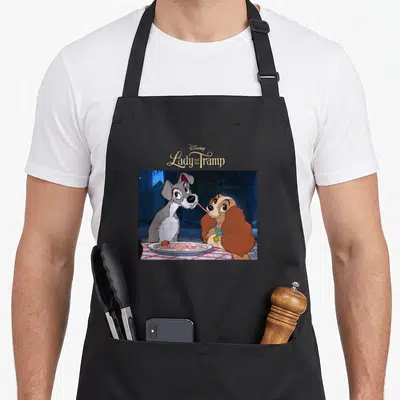 Scene lady and the tramp apron - Disney logo above two cartoon dogs sharing spaghetti on a plate — shown on a black cotton canvas apron against a white background
