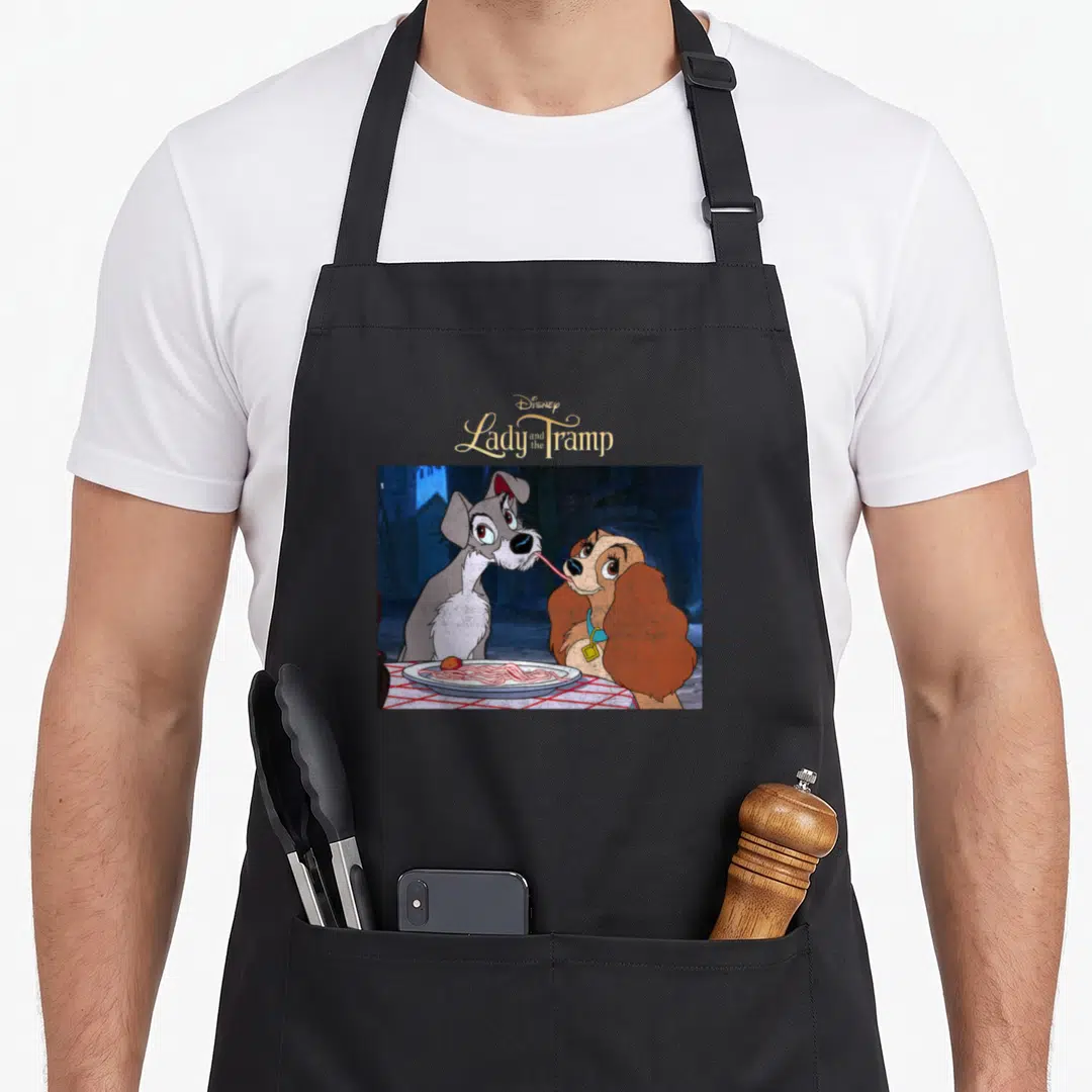 Scene lady and the tramp apron - Disney logo above two cartoon dogs sharing spaghetti on a plate — shown on a black cotton canvas apron against a white background