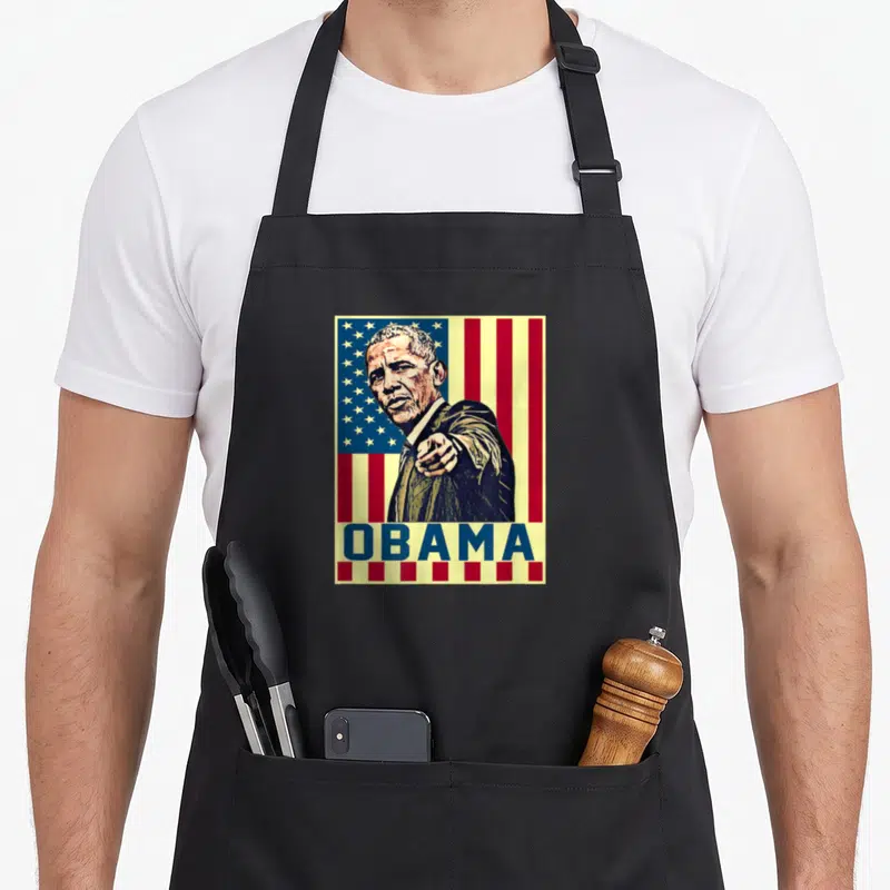 Barack novelty apron - Retro portrait of Obama pointing forward over USA flag with blue OBAMA text bar — shown on a black cotton canvas apron against a white background