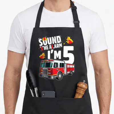 Fire 5th truck apron - SOUND THE ALARM I'M 5 text with fire truck, helmet, extinguisher, and flames — shown on a black cotton canvas apron against a white background