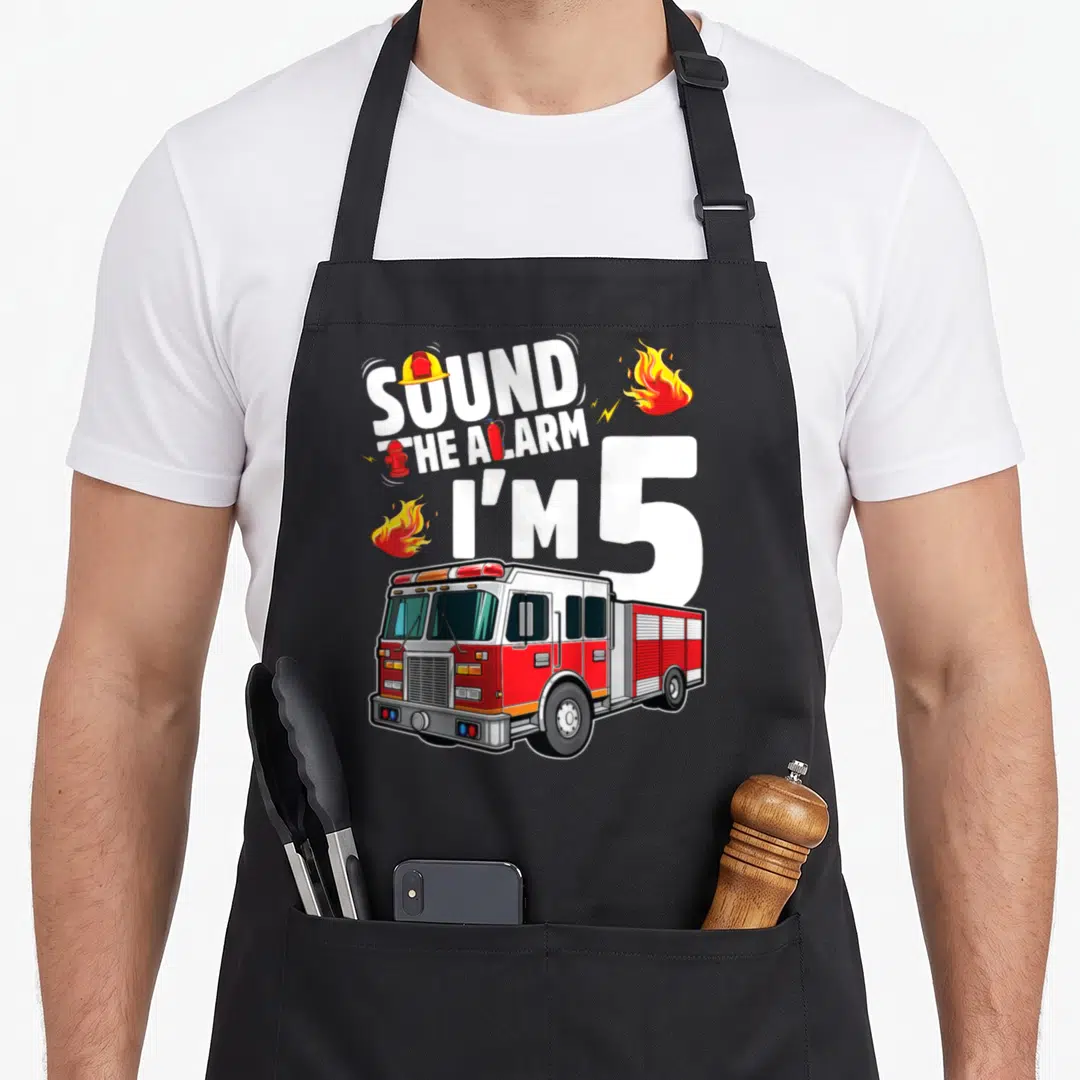 Fire 5th truck apron - SOUND THE ALARM I'M 5 text with fire truck, helmet, extinguisher, and flames — shown on a black cotton canvas apron against a white background
