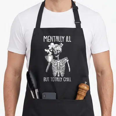 Mentally halloween apron - White distressed skeleton holding steaming mug with sarcastic text — shown on a black cotton canvas apron against a white background