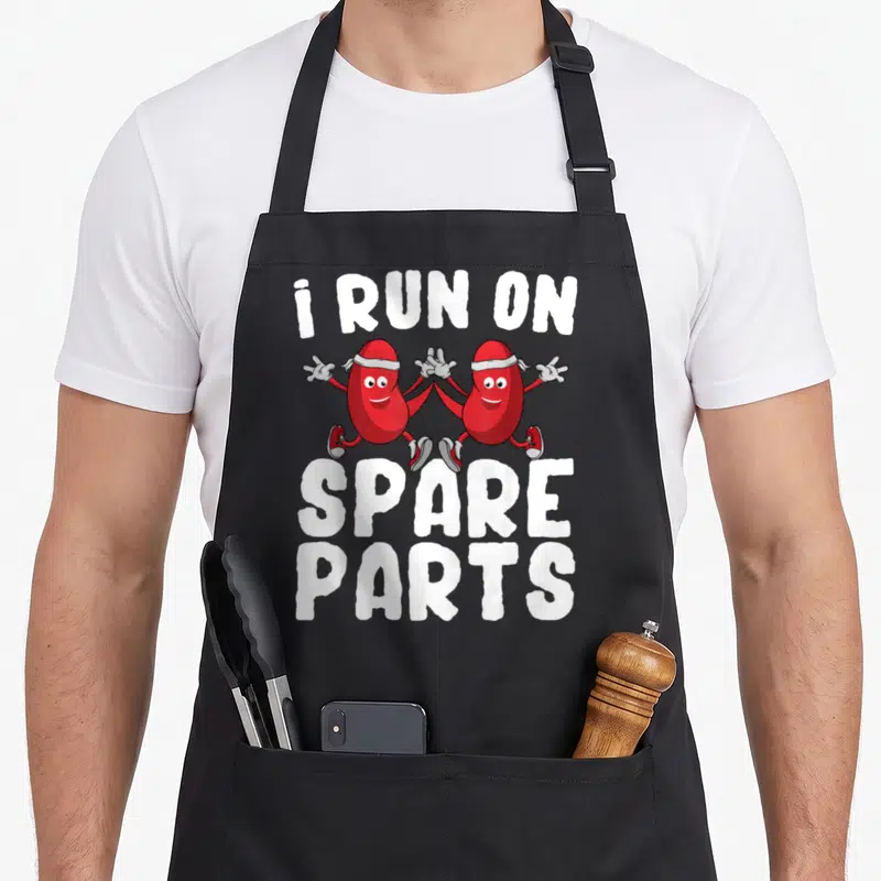 Kidney donor donation organ apron - Two red cartoon kidneys running with white text i RUN ON SPARE PARTS — shown on a black cotton canvas apron against a white background