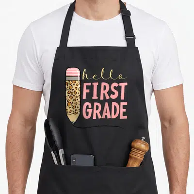 School grade hello apron - Cartoon pencil with leopard print and hello FIRST GRADE text in pink and beige — shown on a black cotton canvas apron against a white background