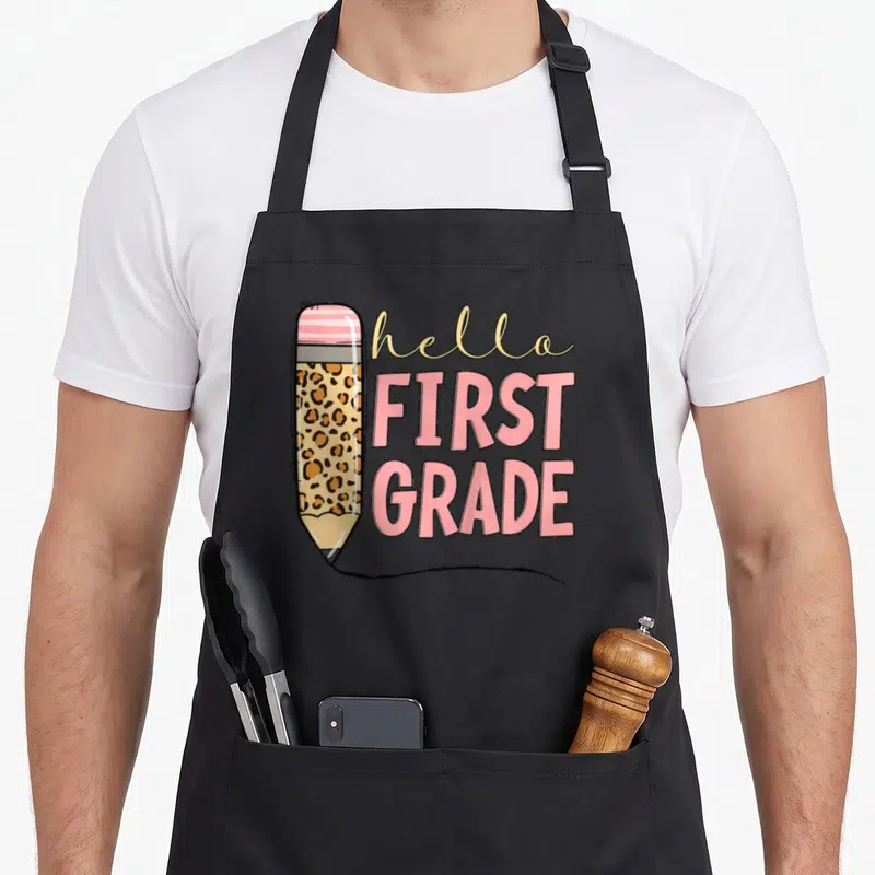 School grade hello apron - Cartoon pencil with leopard print and hello FIRST GRADE text in pink and beige — shown on a black cotton canvas apron against a white background