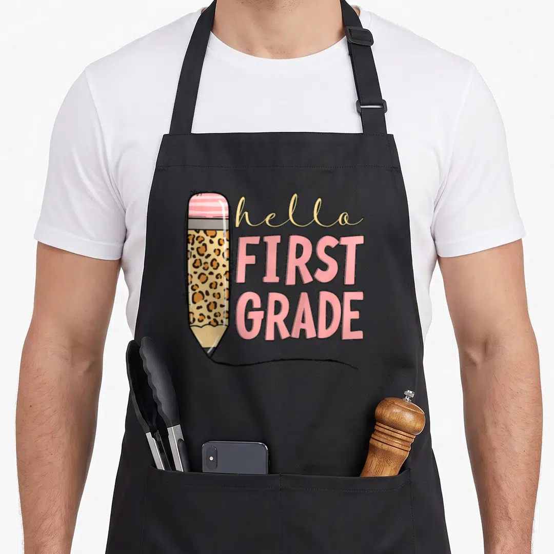 School grade hello apron - Cartoon pencil with leopard print and hello FIRST GRADE text in pink and beige — shown on a black cotton canvas apron against a white background
