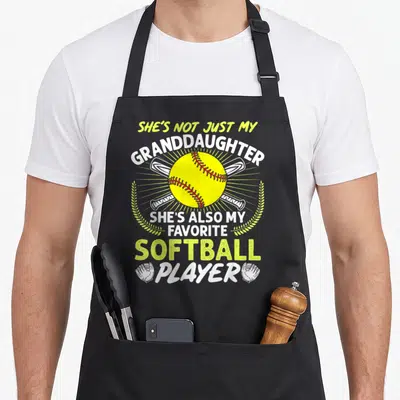 Granddaughter goddaughter apron - Yellow softball with crossed bats and text She's Not Just My Granddaughter She's Also — shown on a black cotton canvas apron against a white background