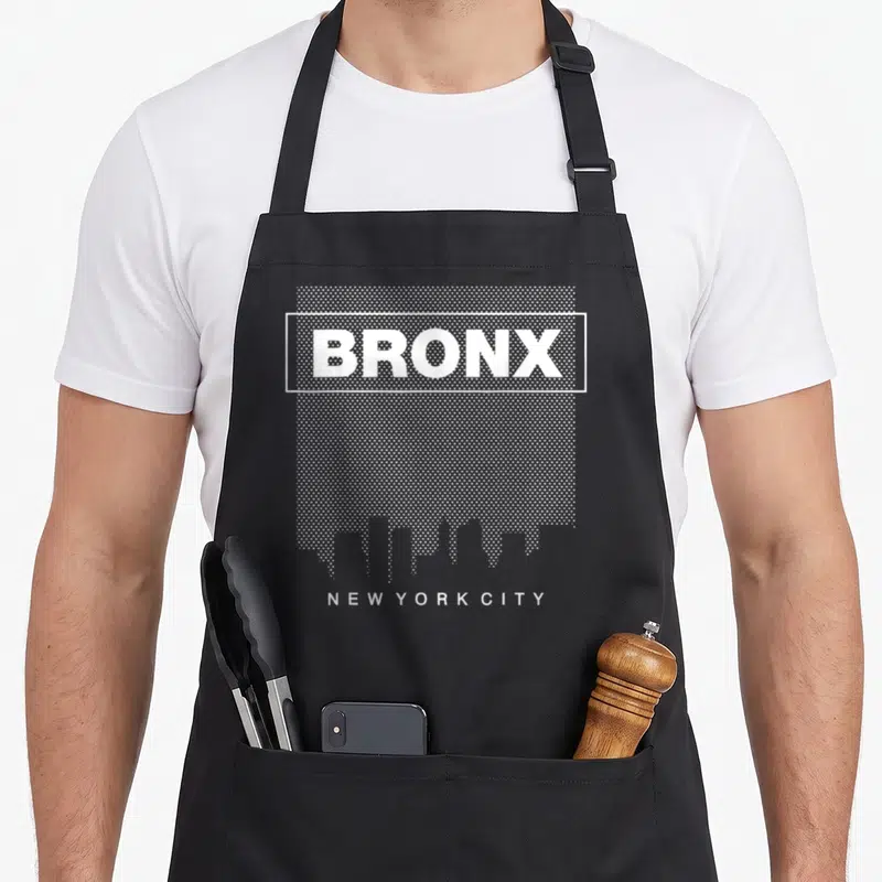 Bronx new york apron for women - White BRONX text in rectangle over dots with skyline and NEW YORK CITY below — shown on a black cotton canvas apron against a white background