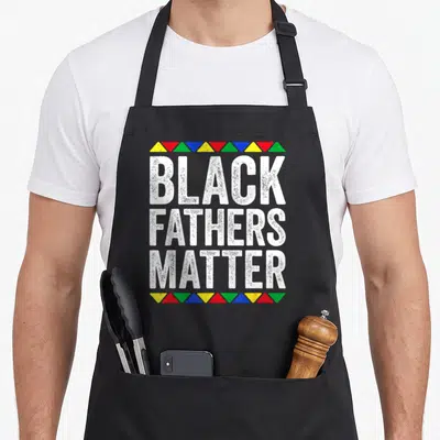 Father black history apron - Black Fathers Matter text framed by rows of red, yellow, blue, green, and white triangles — shown on a black cotton canvas apron against a white background