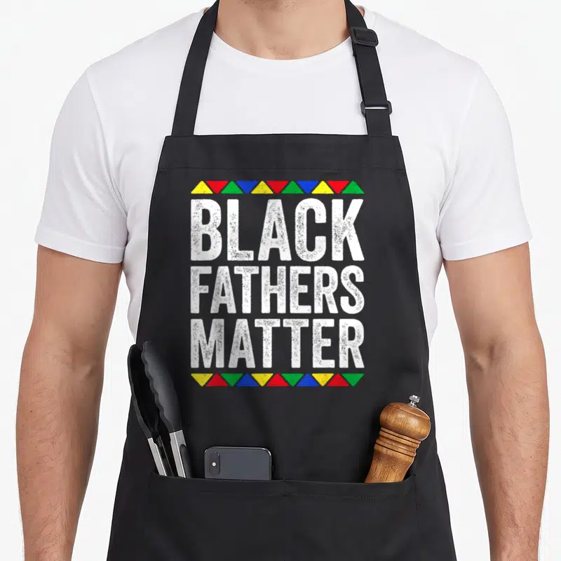 Father black history apron - Black Fathers Matter text framed by rows of red, yellow, blue, green, and white triangles — shown on a black cotton canvas apron against a white background