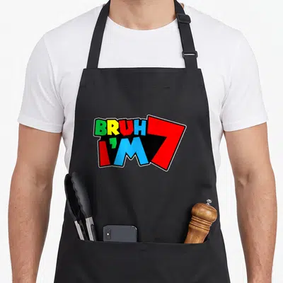 Seven 7yr birthday apron - BRUH I'M 7 text in green yellow red blue black block letters on black background — shown on a black cotton canvas apron against a white background