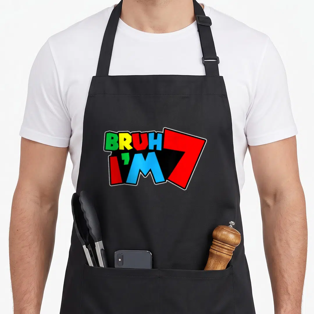 Seven 7yr birthday apron - BRUH I'M 7 text in green yellow red blue black block letters on black background — shown on a black cotton canvas apron against a white background