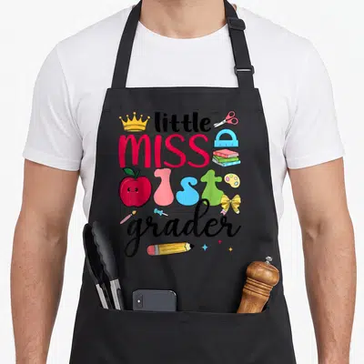 Miss day grade apron - Cartoon text little MISS 1st grader surrounded by an apple, crown, scissors, notebooks, palette — shown on a black cotton canvas apron against a white background