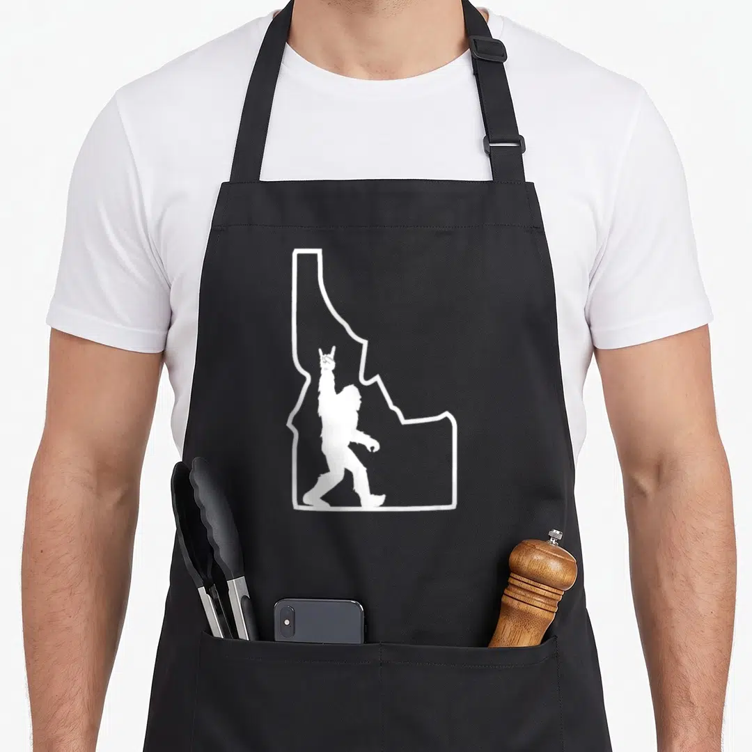 Roll silhouette bigfoot apron - White Bigfoot silhouette raising a hand inside a white Idaho state outline — shown on a black cotton canvas apron against a white background