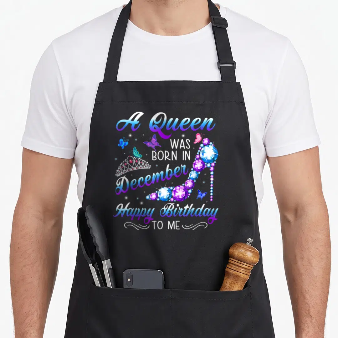 Retro queen birthday apron - Gem covered high heel shoe with crown, butterflies, and December birthday text in blue — shown on a black cotton canvas apron against a white background