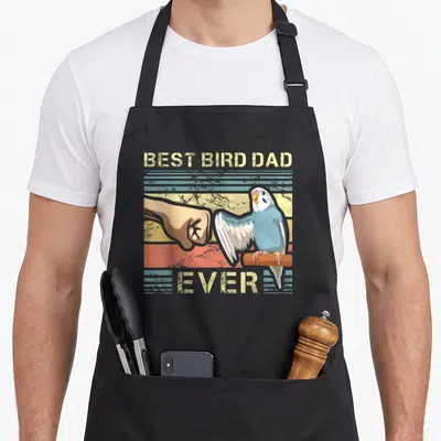 Budgie birds apron - Blue budgie fist bumps human hand under BEST BIRD DAD EVER text on striped background — shown on a black cotton canvas apron against a white background