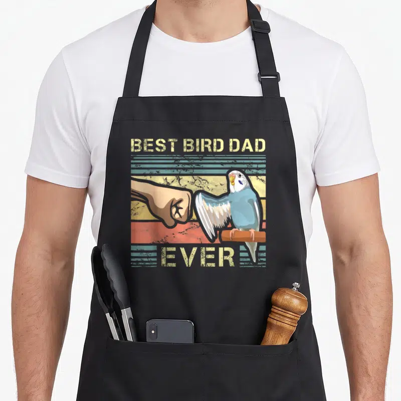 Budgie birds apron - Blue budgie fist bumps human hand under BEST BIRD DAD EVER text on striped background — shown on a black cotton canvas apron against a white background