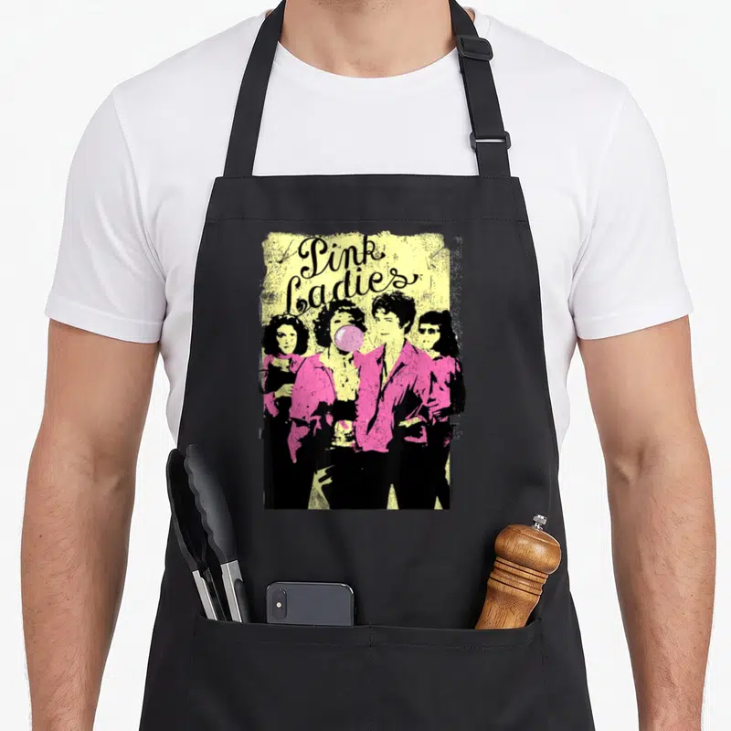 Funny grease apron - Pink Ladies stacked text above four women in pink jackets on yellow distressed background — shown on a black cotton canvas apron against a white background