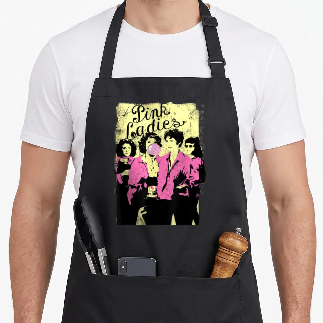 Funny grease apron - Pink Ladies stacked text above four women in pink jackets on yellow distressed background — shown on a black cotton canvas apron against a white background