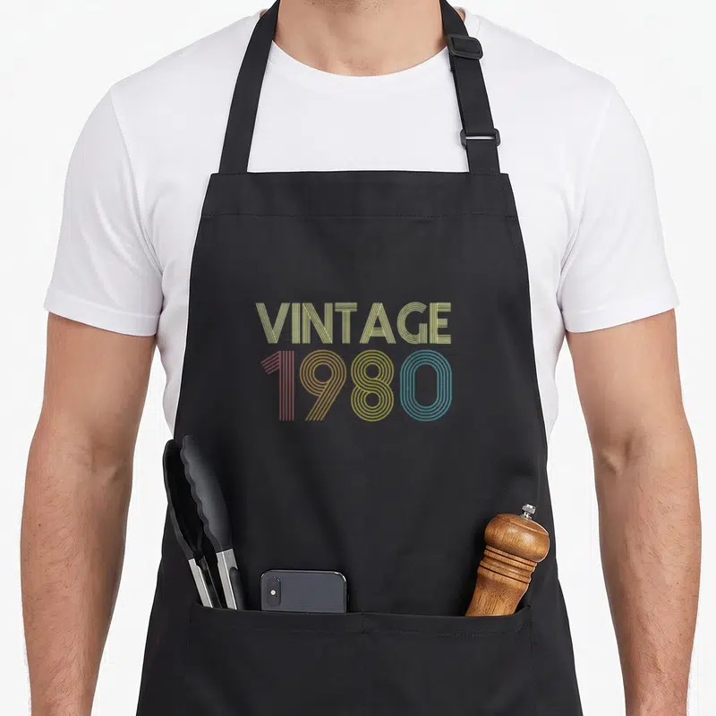 Funny bday 40th apron - VINTAGE 1980 striped typography in yellow red and blue stacked text — shown on a black cotton canvas apron against a white background