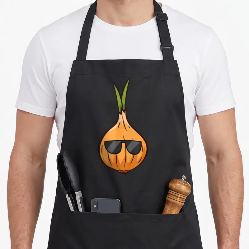 Onion vegetable apron - Cartoon orange onion wearing black sunglasses with green sprouts on top — shown on a black cotton canvas apron against a white background