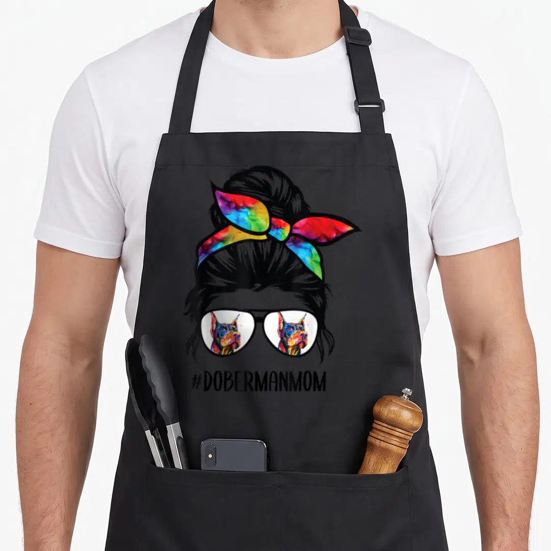 Doberman messy aprons - Woman silhouette with bun, rainbow headband, sunglasses reflecting two dobermans, #DOBERMANMOM — shown on a black cotton canvas apron against a white background