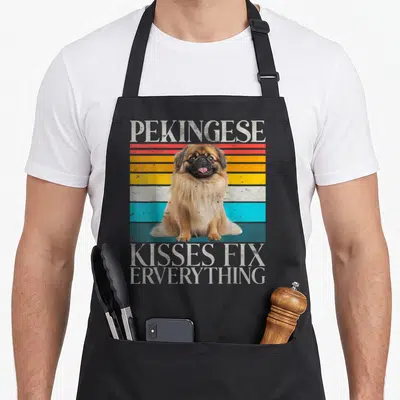 Kiss pekingese apron - Retro Pekingese dog illustration on red, yellow, white, and blue stripes with distressed text — shown on a black cotton canvas apron against a white background