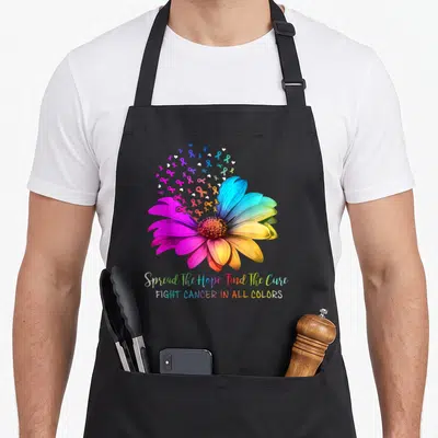 Cancer find aprons - Rainbow flower with multicolored ribbons and white hearts above rainbow text reading Spread — shown on a black cotton canvas apron against a white background