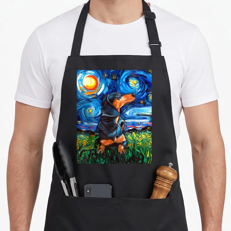 Doxie starry dog apron - Black and tan dachshund sitting in green grass under a swirling blue starry night sky — shown on a black cotton canvas apron against a white background