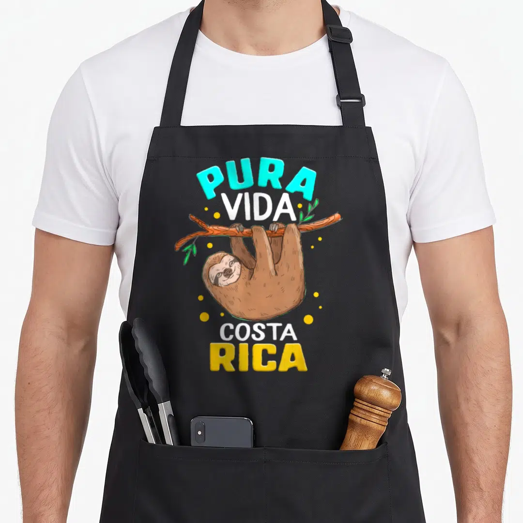Pura sloth apron - Cartoon brown sloth hanging from branch with Pura Vida Costa Rica text in cyan and yellow circles — shown on a black cotton canvas apron against a white background
