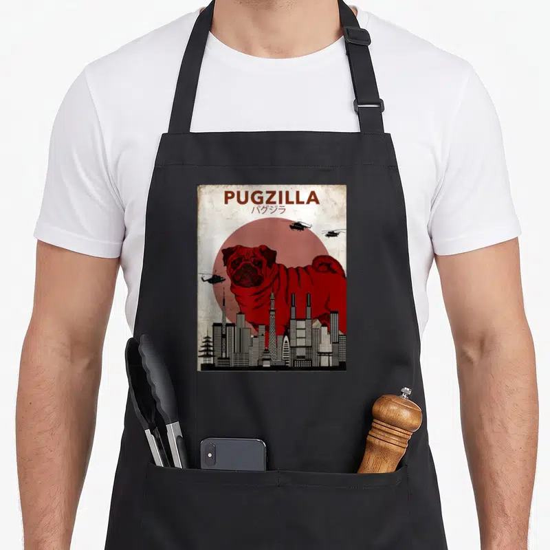 Pug cartoon apron - Red pug monster behind black skyline with PUGZILLA text and three helicopters — shown on a black cotton canvas apron against a white background