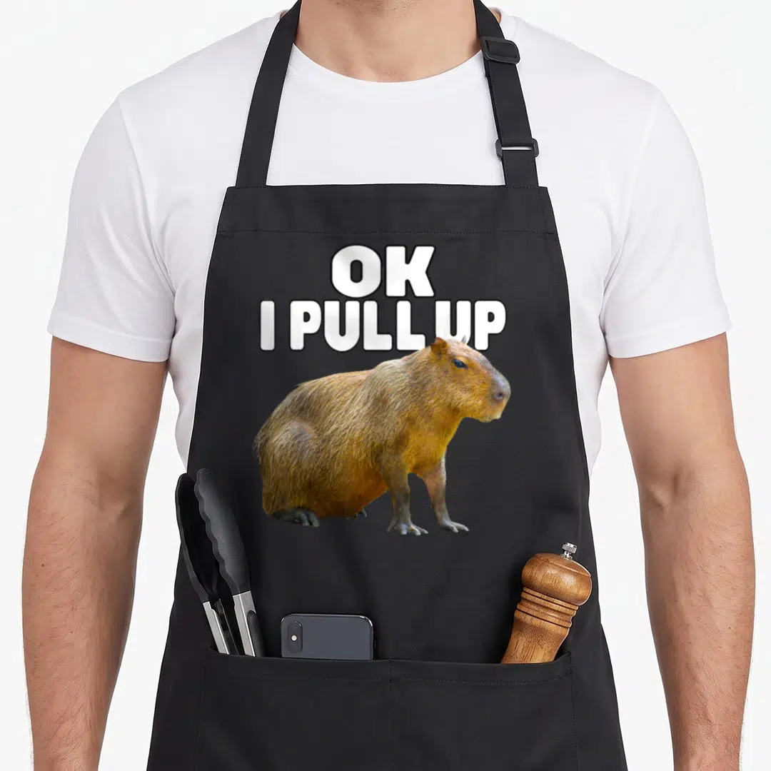 Pull meme apron - Stacked white text OK I PULL UP above a standing brown capybara photo — shown on a black cotton canvas apron against a white background