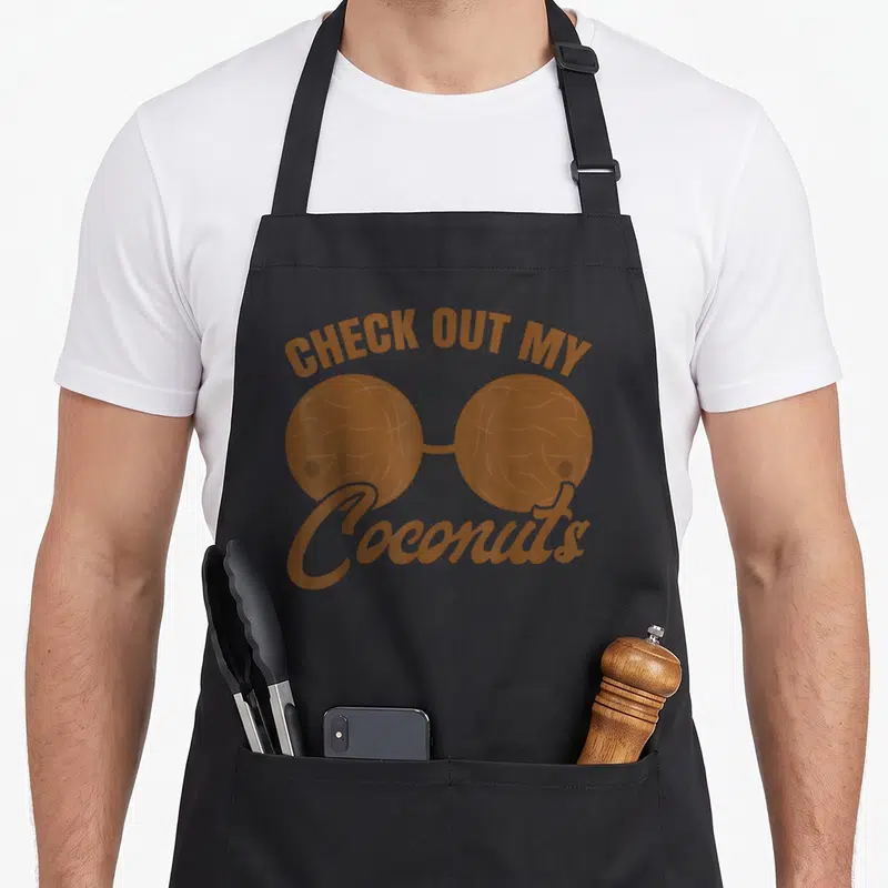 Bra coconut apron - Two brown coconuts connected by a bridge forming a bra shape with arched text above and script below — shown on a black cotton canvas apron against a white background