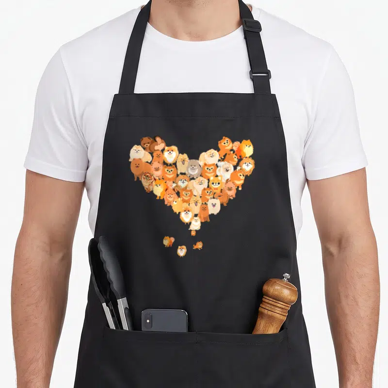 Retro pomeranian dog apron - Cartoon Pomeranians in orange tan white forming a heart shape with three dogs below — shown on a black cotton canvas apron against a white background
