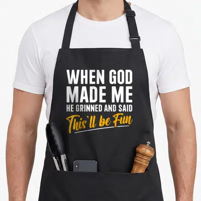 Cute god funny christian apron - White and yellow stacked text reading When God Made Me He Grinned And Said This'll be — shown on a black cotton canvas apron against a white background