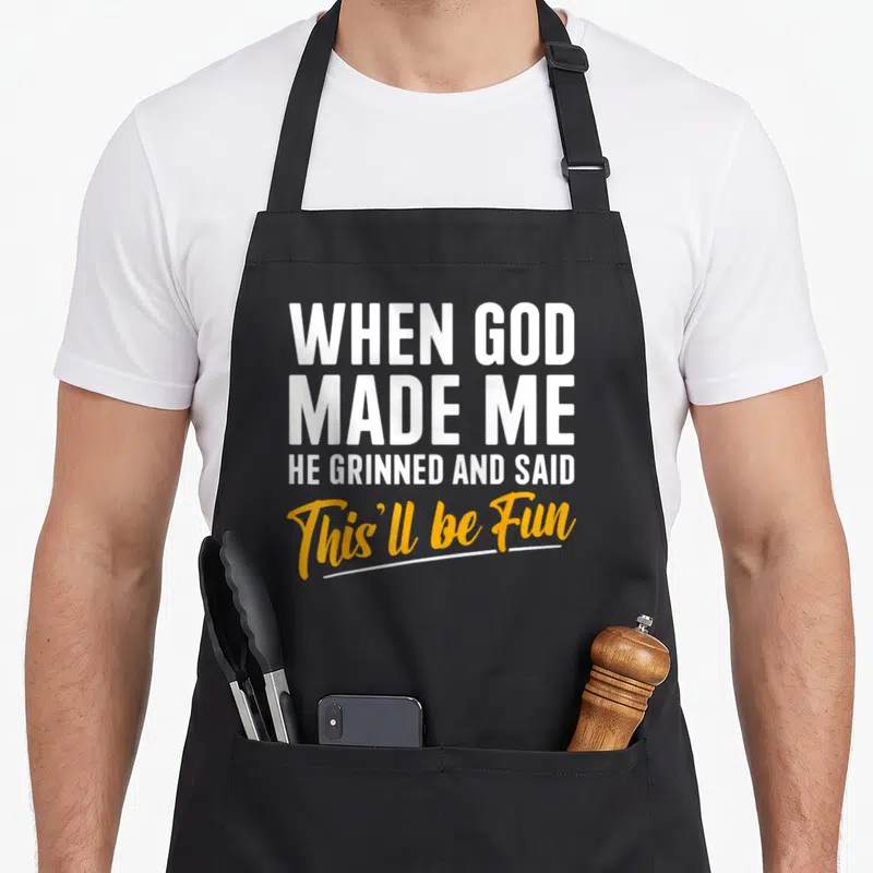 Cute god funny christian apron - White and yellow stacked text reading When God Made Me He Grinned And Said This'll be — shown on a black cotton canvas apron against a white background