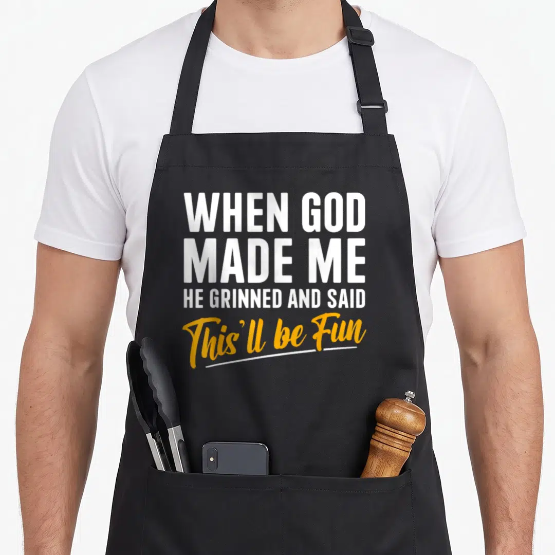 Cute god funny christian apron - White and yellow stacked text reading When God Made Me He Grinned And Said This'll be — shown on a black cotton canvas apron against a white background