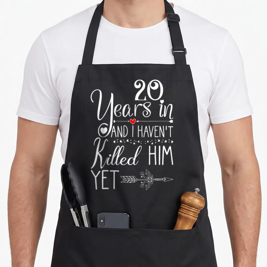 20th wife apron - White stacked text 20 Years in and I haven't killed him yet with red and white hearts — shown on a black cotton canvas apron against a white background