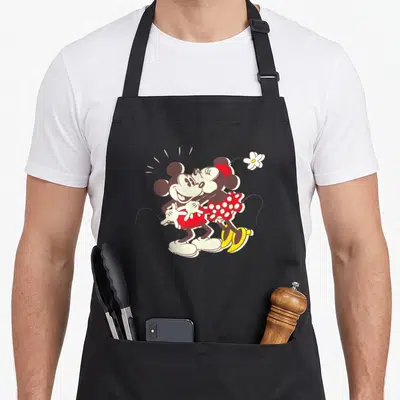 Kiss i love my mouse apron - Mickey and Minnie Mouse embracing in vintage cartoon style with flower and motion lines — shown on a black cotton canvas apron against a white background