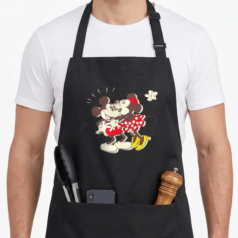 Kiss i love my mouse apron - Mickey and Minnie Mouse embracing in vintage cartoon style with flower and motion lines — shown on a black cotton canvas apron against a white background